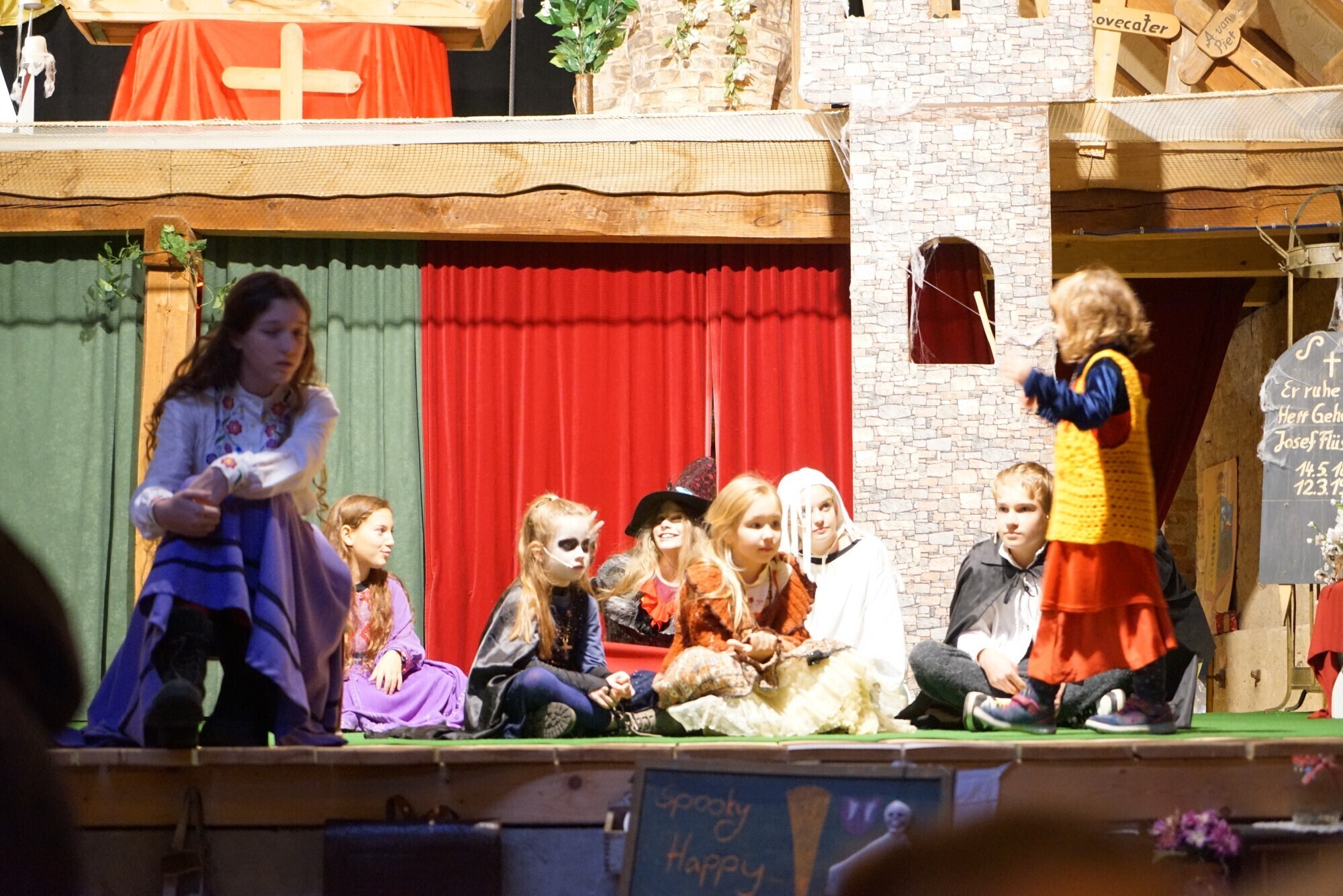 Zur vollständigen Stück-Galerie wechseln Kinder auf der Bühne beim Halloween-Kürbisfest. Kinder in bunten Kostümen spielen Szenen des Märchen-Musicals „Kürbisfest und Maskenball mit Graf Dracula“ auf der Bühne, mit Vampir- und Hexenmasken vor Burgkulisse.